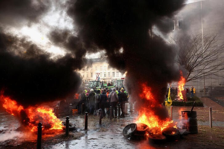 Farmers’ Anger Spreads In Europe, Governments Promise Help Smoke rises from objects set on fire as people gather in front of the European Union headquarters as Belgian farmers use their tractors to block the headquarters