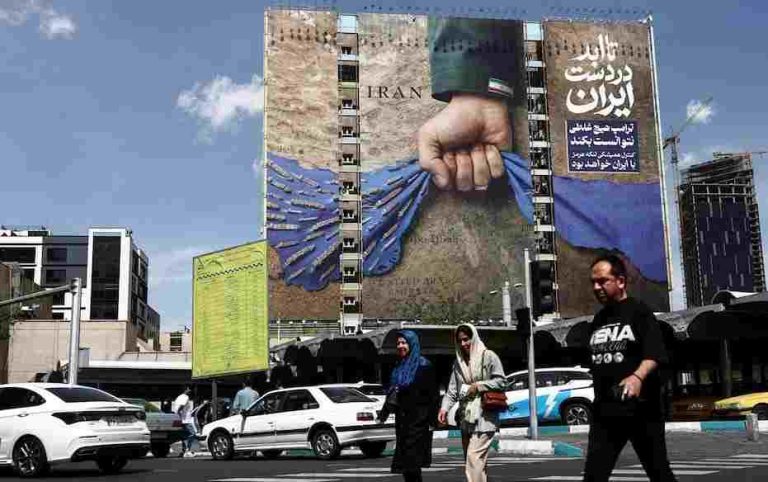 People walk past a billboard with a graphic design about the Strait of Hormuz on a building.