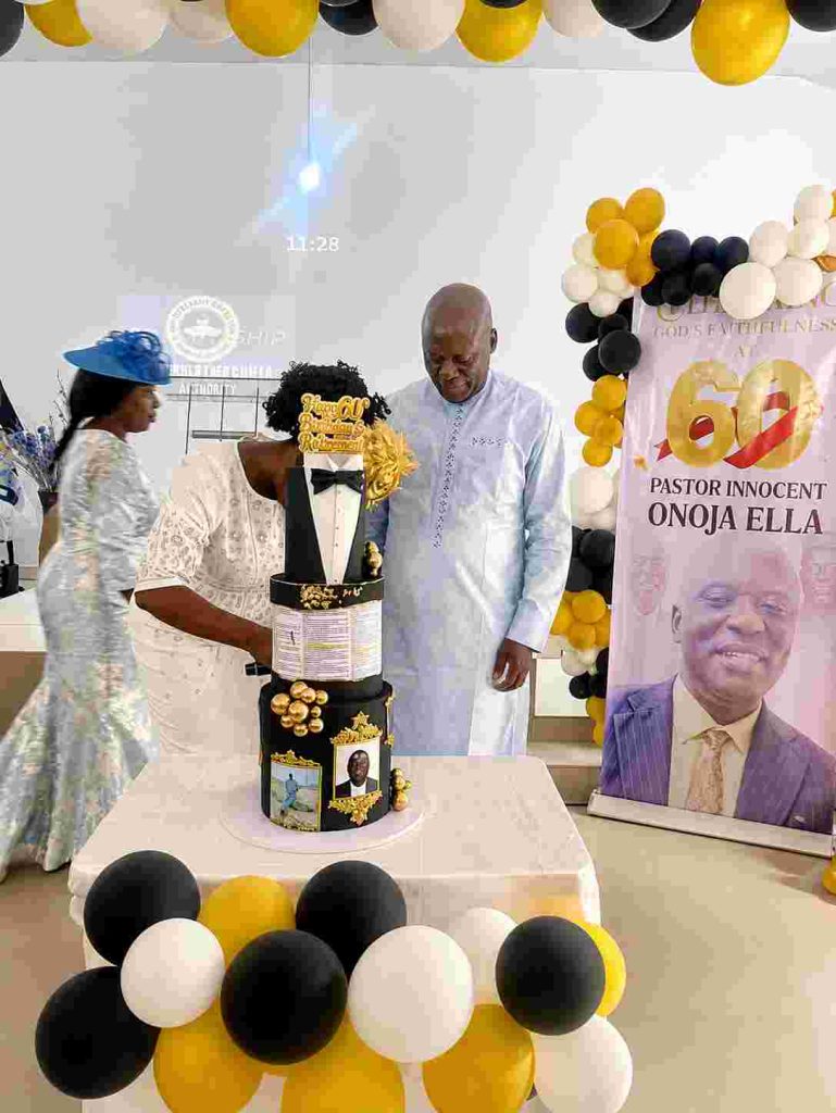 Onoja standing before the birthday cake, as his wife moves to join him.